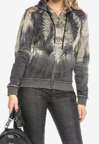 Hoodie met ritssluiting in vervaagd grijs met zwarte arendgrafiek, rafelige zoom en verweerde manchetten. Gecombineerd met glanzende zwarte textuurspijkerbroek en handtas.