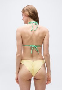 Γυναίκα με μακριά ανοιχτό καστανά μαλλιά, που φοράει πράσινο δέσιμο πάνω μέρος μπικίνι και κίτρινο κάτω μέρος μπικίνι, βλέπεται από πίσω σε απλό φόντο.