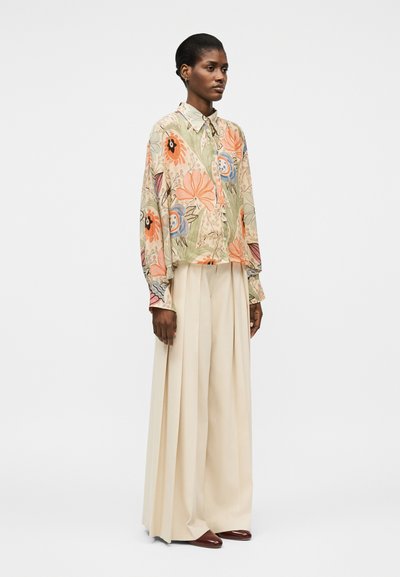 Femme debout portant un chemisier à manches longues à fleurs et un pantalon large crème avec des chaussures marron, faisant face légèrement à droite sur un fond uni.