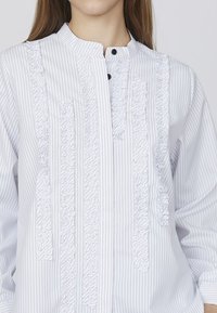 Hvidstripet skjorte med knapper, der har teksturerede flæser langs frontpladen og en krave i båndstil, med subtile sorte knapdetaljer.