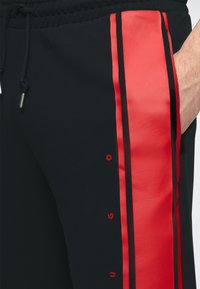 Gros plan d’un pantalon noir avec des rayures verticales rouges et de petites lettres rouges le long de la rayure, main partiellement dans la poche, et cordon noir visible.