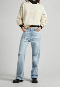 Lichtbeige sweatshirt met reliëfbranding, gecombineerd met lichtblauwe, wijde jeans met een versleten knie. Zwarte enkellaarsjes maken de look af.