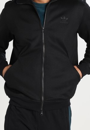 Person trägt eine schwarze Adidas Zip-Up-Jacke mit den Händen in den Taschen und zeigt das Logo auf der linken Brust vor einem schlichten Hintergrund.