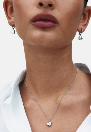 Hõbedast südamekujuga kõrvarõngad ja kaelakee modellil, peene kettaga ja poleeritud viimistlusega hele tausta ees.