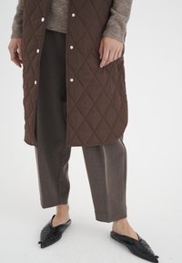 Brun quiltad lång väst med silverfärgade tryckknappar, lagd över en ljusbrun strukturerad tröja och mörkbruna skräddarsydda byxor, ihop med svarta ballerinaskor.