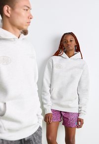 To mennesker bærer lysegrå hættetrøjer; den ene vender fremad iført pink leopardmønstret shorts, mens den anden er sløret i profil mod en hvid baggrund.