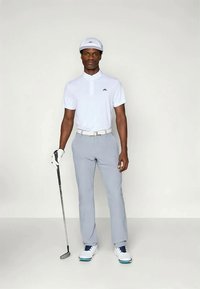 Valkoinen golfpaita, jossa on kaulus ja logo, vaaleanharmaat housut, valkoiset golfkengät sinisillä yksityiskohdilla, valkoinen lippalakki, pitelee golfmailaa.