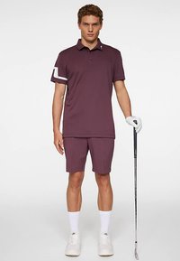 Tummanpunainen poolopaita, jossa on valkoinen yksityiskohta hihassa, yhdistettynä samanvärisiin shortseihin. Malli pitää kädessään golfmailaa ja käyttää valkoisia kenkiä ja sukkia.