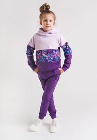 Fioletowy dres z liliowym, z kapturem, z akcentami w kwiatowy wzór oraz dopasowanymi fioletowymi spodniami. Stylizację uzupełniają białe trampki.