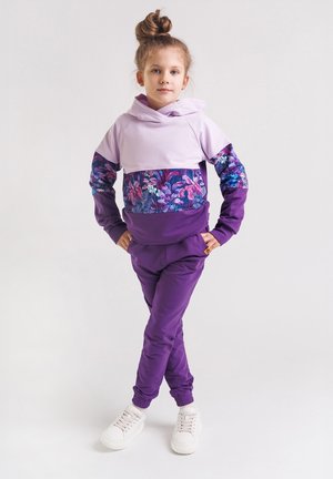 Fioletowy dres z liliowym, z kapturem, z akcentami w kwiatowy wzór oraz dopasowanymi fioletowymi spodniami. Stylizację uzupełniają białe trampki.