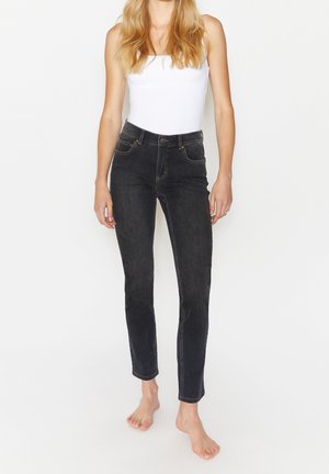 Femme debout pieds nus portant un haut blanc sans manches et un jean noir taille haute devant un fond blanc uni.