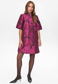 Ženska, oblečena v temno vijolično obleko z velikimi svetlo rožnatimi cvetličnimi motivi, črnimi prosojnimi najlonkami in črno koničastimi slingback petkami.