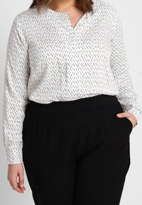 Chemisier blanc à manches longues avec des lignes verticales noires, col en V, porté avec un pantalon noir taille haute avec des plis à l'avant et des poches.