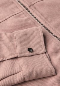 Detailní záběr rukávu trubkového kabátu v jemné růžové barvě s černým knoflíkem a blízkým stříbrným zipem na jemném textilním pozadí.