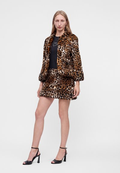 Femme portant une veste en fausse fourrure imprimé léopard et une jupe assortie, un top noir et des sandales noires à talons hauts, debout sur un fond blanc.
