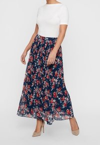 Kvinna som bär en vit kortärmad topp och en marinblå veckad maxikjol med röd och vit blommönster, parat med beige klackar.