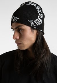 Bonnet en tricot noir avec un texte blanc entourant le bord. Le matériau semble doux, avec un ajustement serré et une texture lisse.