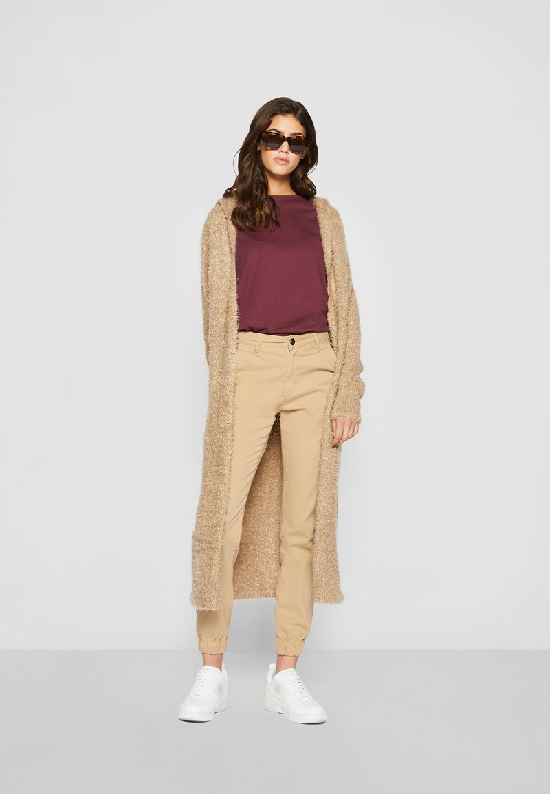 Lang beige fluffy cardigan over en maroon t-skjorte, kombinert med beige bukser og hvite joggesko. Modellen har på seg solbriller.