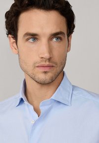 Camisa de botón azul claro con cuello puntiagudo, hecha de una tela suave, que presenta una textura sutil y un solo botón en el cuello.