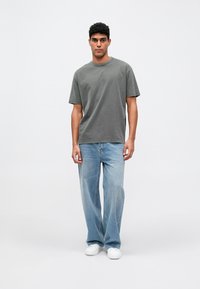 Muž stojaci v tlmenom zelenom oversized tričku, vo voľných svetlých modrých džínsach a bielych teniskách proti jednoduchej bielej pozadí.