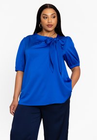 Blouse bleu royal à manches bouffantes courtes et détail de nœud noué au niveau du col. Tissu lisse et brillant avec une coupe décontractée.