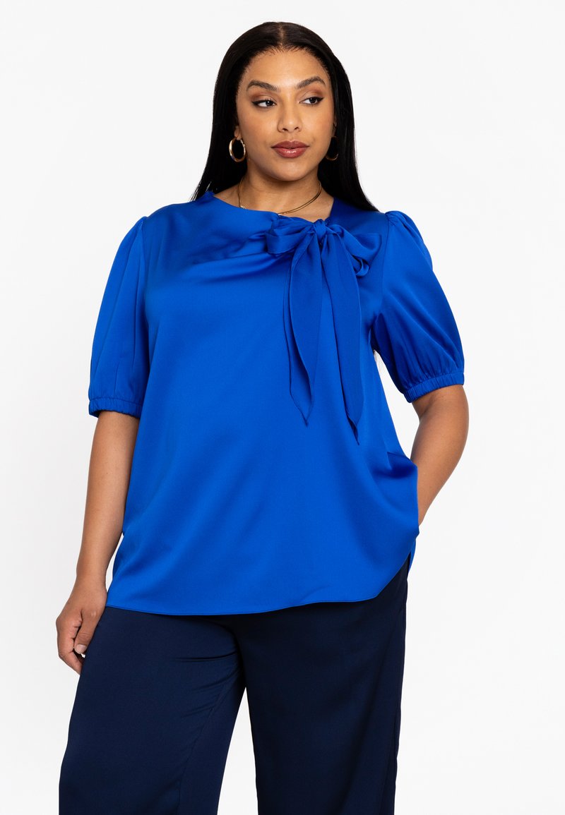 Blouse bleu royal à manches bouffantes courtes et détail de nœud noué au niveau du col. Tissu lisse et brillant avec une coupe décontractée.