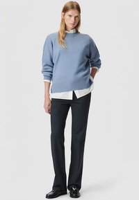 Hellblauer Strickpullover mit hohem Kragen, überschnittenen Schultern, kombiniert mit weißem Hemd und dunkler, maßgeschneiderter Hose, ergänzt durch schwarze Schuhe.