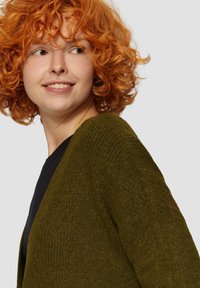 Olivgrüner gestrickter Cardigan mit strukturiertem Muster, offenem Frontdesign und gerippten Details entlang der Kanten, getragen über einem schwarzen Oberteil.