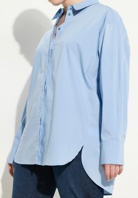 Camicia azzurra chiara con colletto strutturato, maniche lunghe, orlo asimmetrico e tessuto morbido. Presenta bottoni a contrasto.