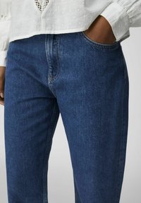 Person som bär blå denimjeans och en vit strukturerad långärmad blus, med en hand i jeansfickan mot en plain bakgrund.