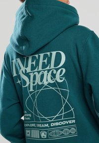 Tealitakki, jossa on suuri valkoinen teksti "Tarvitsen avaruutta" ja graafisia kuvastoja, mukaan lukien pyöreitä kuvioita ja avaruuskypäräikoni.