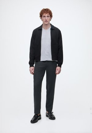 Νέος άνδρας με σγουρά κόκκινα μαλλιά, ντυμένος με μαύρο μπουφάν, ανοιχτό γκρι μπλουζάκι με V λαιμόκοψη, σκούρο παντελόνι και μαύρα δερμάτινα παπούτσια, στέκεται μπροστά σε λευκό φόντο.