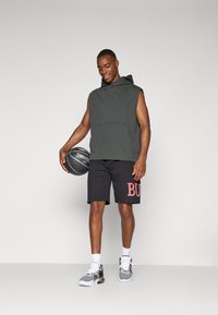 Sudadera sin mangas de color verde oscuro con un bolsillo frontal y un logo, combinada con pantalones cortos negros que tienen texto rojo. Sostiene un balón de baloncesto y lleva calzado deportivo.