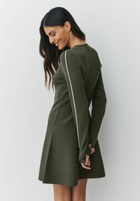 Olijfgroene geribbelde jurk met lange mouwen, met een aansluitend lijfje en een uitlopende rok, verfraaid met witte strepen langs de mouwen.