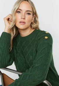 Zielony sweter z dzianiny z wzorem w warkocze, z złotymi guzikami na ramionach i ściągaczami na rękawach. Miękka tekstura.