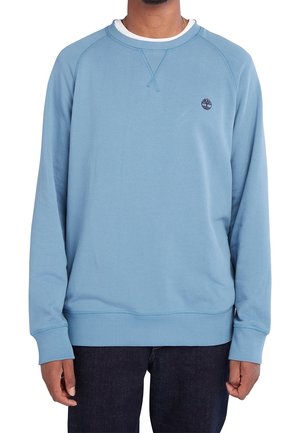 Muž v světle modré mikině s dlouhým rukávem a malým tmavým logem na levé straně hrudi, tmavé kalhoty, stojící před bílým pozadím.