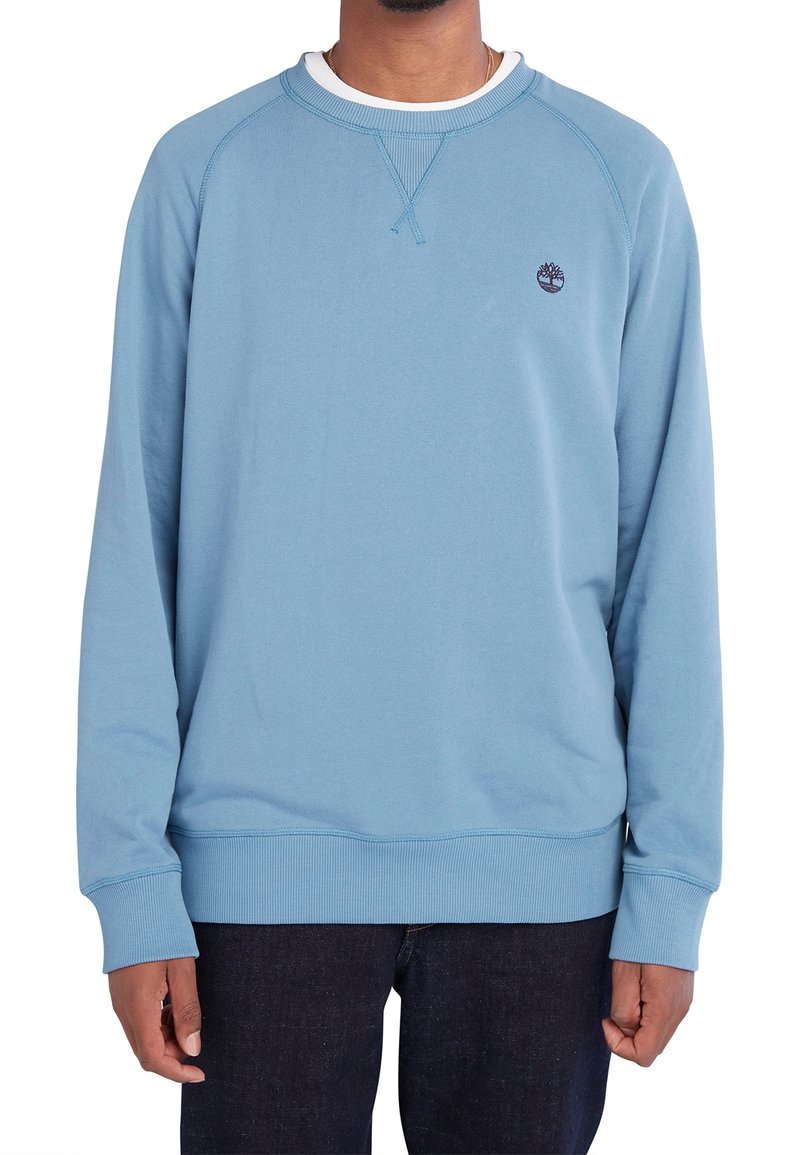 Homme portant un sweat-shirt à manches longues bleu clair avec un petit logo sombre sur le côté gauche de la poitrine et un pantalon sombre, debout devant un fond blanc.