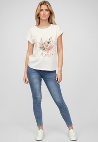 T-shirt crème à manches courtes avec un design graphique floral et de plumes, associé à un jean skinny bleu et des baskets blanches. Texture de tissu lisse.
