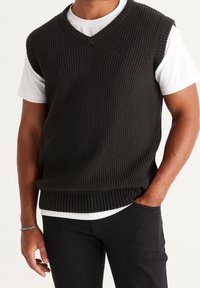 Maglione smanicato lavorato a maglia di colore verde scuro con scollatura a V profonda e motivo a costine, indossato sopra una t-shirt bianca a maniche corte. Visibili pantaloni neri.