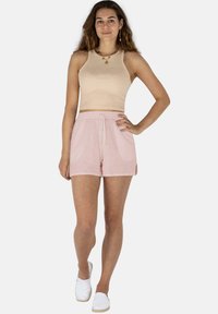 Frau steht mit einer Hand auf der Hüfte und trägt ein beiges ärmelloses Crop-Top, rosa Shorts mit Kordelzug, weiße Slipper und mehrschichtige Halsketten.