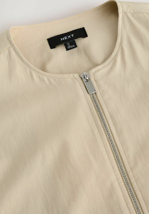 Beige jacka med rund halsringning och en silvrig dragkedja. Tillverkad av glatt tyg med en subtil lyster. Lappen säger "NEXT".