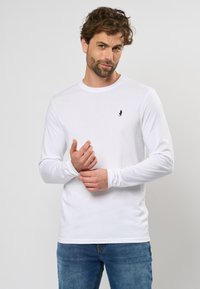 Camicia bianca a maniche lunghe in cotone, con collo tondo e un piccolo logo blu navy sul petto sinistro. Abbinata a jeans in denim blu.