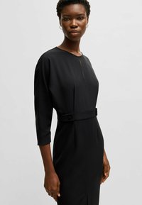 Svart klänning med rund halsringning, trekvarts ärmar och figursydd midja med en knappdetalj. Slätt material med minimala veck.