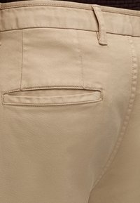 Poche arrière beige du pantalon avec passants de ceinture et surpiqûres visibles sur tissu sergé.