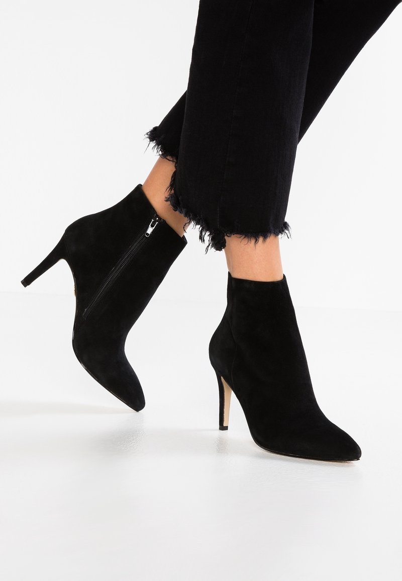 Bottines en daim noir à bout pointu, avec fermetures éclair latérales et talons stilettos, portées avec un pantalon noir à ourlet effiloché.
