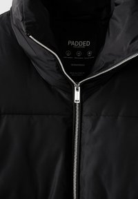 Veste matelassée noire avec fermeture éclair argentée, col montant et étiquette indiquant des propriétés déperlantes, résistantes au vent et d'isolation thermique.