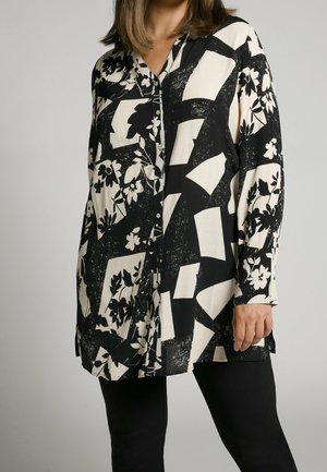 Kobieta w czarno-białej bluzce na guziki z długim rękawem, ozdobionej kwiatowym i abstrakcyjnym wzorem, oraz w czarnych spodniach na jednolitym tle.