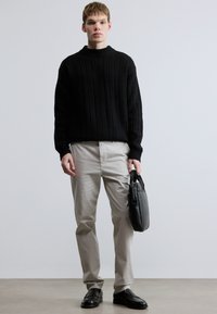 Sort ribstrikket sweater parret med beige chinos. Modellen holder en sort lædertaske og har sorte slip-on sko på. Neutralt baggrund.