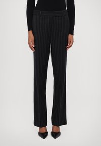 Un pantalon noir à rayures fines au design ajusté, présentant une coupe droite et une texture de tissu lisse, associé à des chaussures noires à bouts pointus.