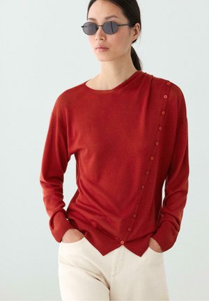 Donna con occhiali da sole rotondi neri, maglione rosso con bottoni diagonali e pantaloni beige chiaro, in piedi con le mani nelle tasche.